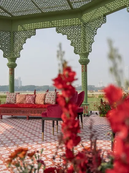 A view from behind the vibrant red flowers lining the aisle, looking towards the couple's seating inside the mandap. This perspective highlights the lushness of the floral design.