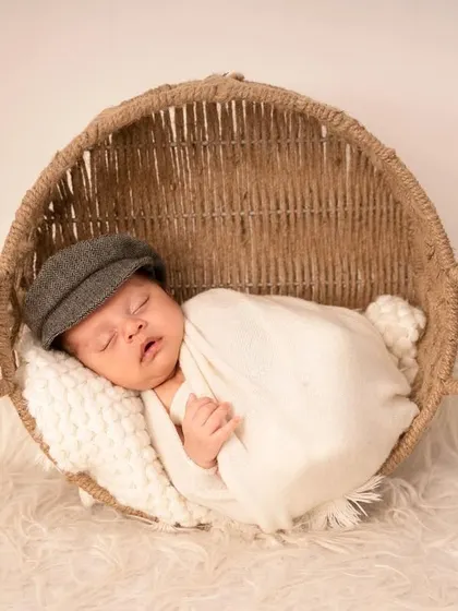 A simple and classic portrait of a baby in a newsboy cap, swaddled in white and resting in a natural fiber basket.