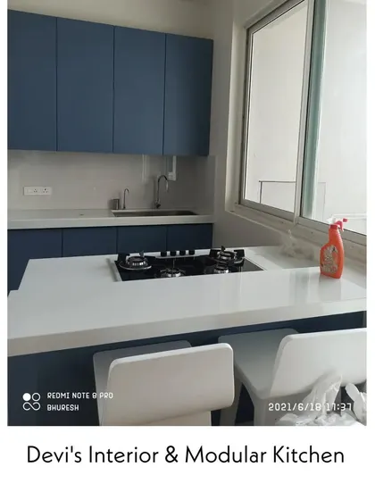 This kitchen in Andheri features a bold blue matte finish and a clean white countertop that extends into a breakfast bar. The large window allows for plenty of natural light, making the space feel open and airy.