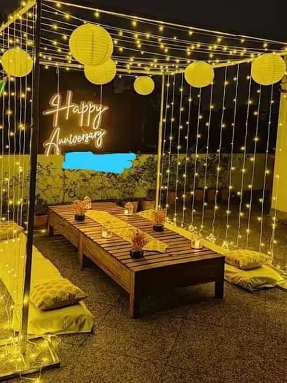 An elegant anniversary celebration on a terrace. This setup features a beautiful cabana draped in fairy lights, paper lanterns, and a neon "Happy Anniversary" sign, with comfortable floor seating for a relaxed party vibe.