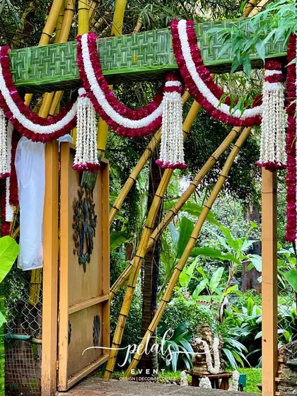 This traditional wedding entrance arch is wrapped in woven coconut leaves and decorated with garlands of maroon and white chrysanthemums, creating a rich, natural texture.