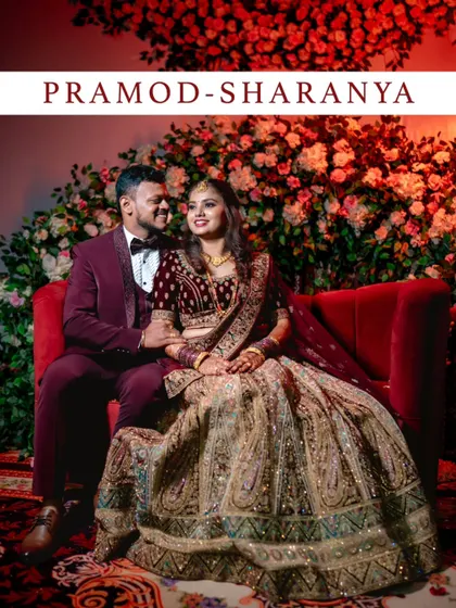 A grand, poster-style portrait of a couple at their reception, seated on a red sofa against a stunning floral backdrop.