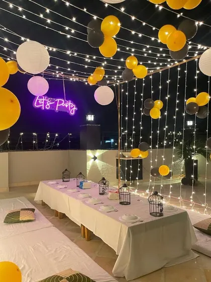 The same terrace party setup at night, with the fairy lights and "Let's Party" neon sign turned on, creating a magical atmosphere.
