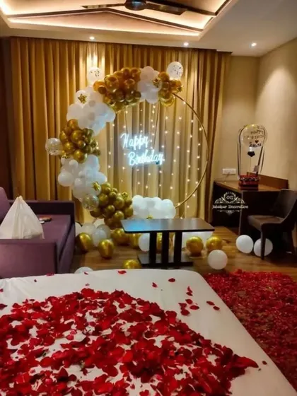 A luxurious hotel room birthday surprise. A gold and white balloon arch with a neon sign is set up by the bed, which is covered in red rose petals for a touch of romance.