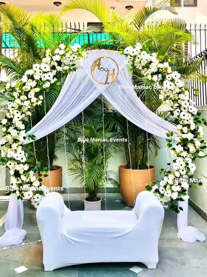 A stunning outdoor day-time setup for a Roka or Nikkah. The circular arch is adorned with white flowers and drapes, beautifully complemented by the natural greenery in the background.