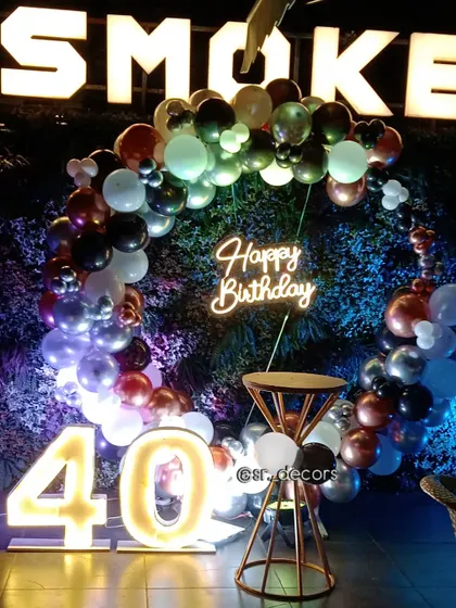 This 40th birthday setup at a restaurant features a multi-colored balloon ring against a green wall, a neon sign, and large marquee numbers.