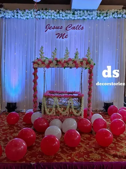 An elegant stage for a naming ceremony or Baptism, featuring a beautifully decorated jhula (cradle). The backdrop of white curtains with blue uplighting creates a serene and divine atmosphere.