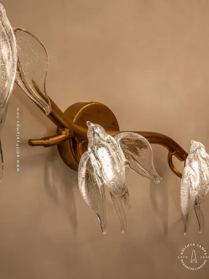 A close-up of our bird-themed wall sconce. Three delicate glass birds are mounted on a small rose-gold branch, providing a soft, ambient light. It's a charming accent piece that complements the larger chandeliers in the series or stands beautifully on its own.