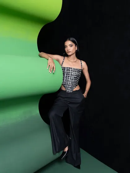 A full-length shot of a single model, showcasing a modern look with a grid-patterned corset and wide-leg trousers. The pose is relaxed yet confident, set against the graphic backdrop.