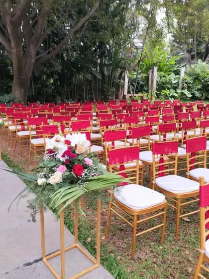 An outdoor ceremony setup with rows of chairs decorated with bright red sashes, creating a bold and vibrant look. A floral arrangement on a stand marks the aisle.