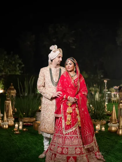 A classic wedding portrait of the couple in their stunning red and cream attire. The elegant outdoor decor with candles creates a magical, romantic atmosphere for this shot.