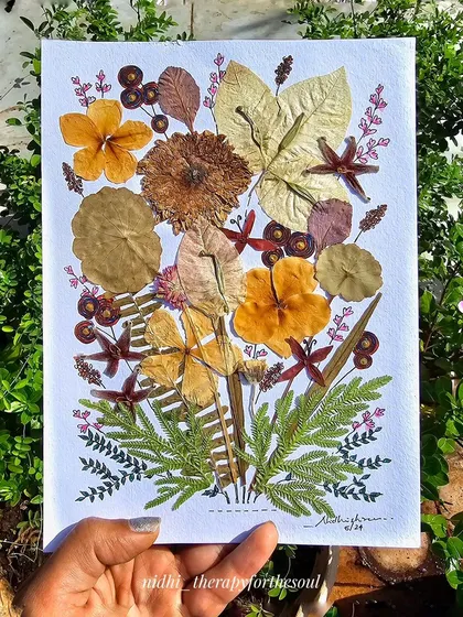 A hand holding the pressed flower art in a garden setting, surrounded by living plants.