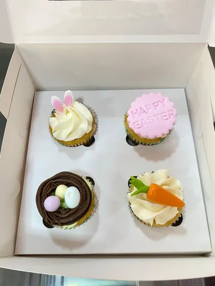 A box of four assorted Easter-themed cupcakes, featuring bunny ears, a bird's nest, a carrot, and a "Happy Easter" message.