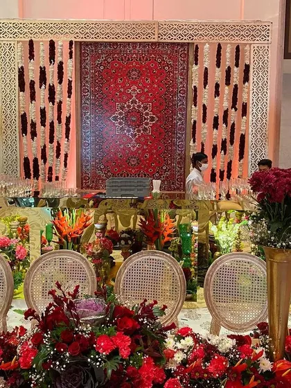 A bar designed for a royal wedding, featuring a stunning backdrop made from a traditional red carpet. The space is overflowing with red floral arrangements and lit with the warm glow of classic Indian lamps.