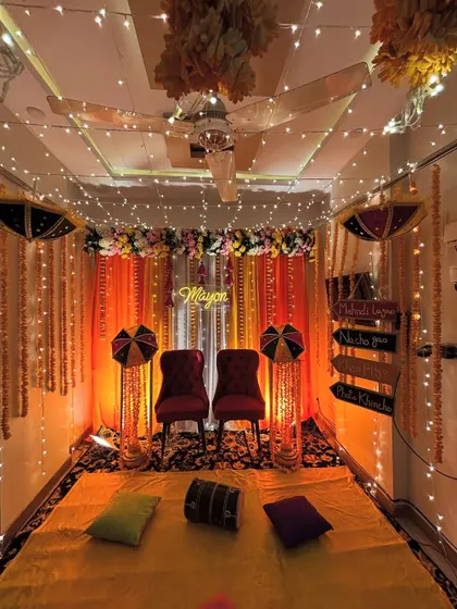 A full view of an indoor Haldi or Mayon setup. The decor features a seating area on the floor with colorful cushions, against a backdrop of orange drapes and marigold garlands.
