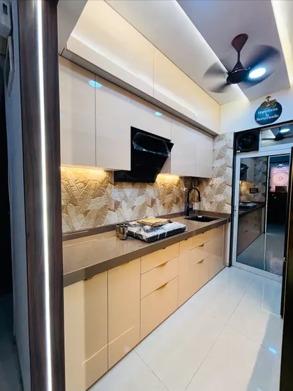 This parallel kitchen design creates a warm and luxurious feel with high-gloss beige cabinets and ambient lighting. Note the textured stone backsplash and integrated black chimney, creating a seamless and elegant workspace.