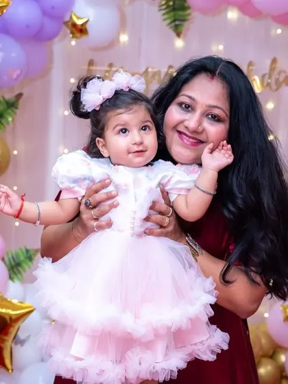 A mother holds her adorable baby girl, both smiling for the camera at a birthday party. We focus on capturing the special bond between parents and their children.