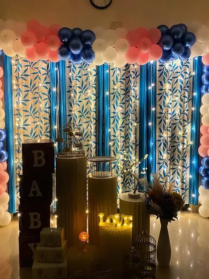 A gender reveal or baby shower setup at home. The balloon arch uses pink, blue, and white balloons, with 'BABY' blocks and other props arranged in front of a curtain with fairy lights.