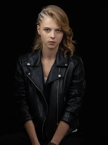 A classic portrait in a leather jacket against a black background. The lighting is soft, focusing on the model's direct and engaging expression.