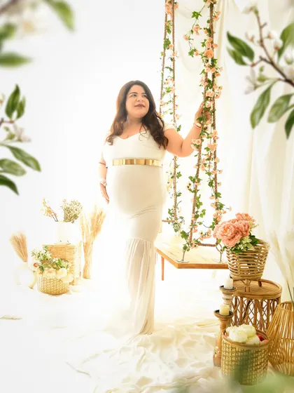 A beautiful solo portrait of the mom-to-be in a white gown, standing by our floral swing. The soft focus on the foreground flowers adds a dreamy, artistic touch to the image.