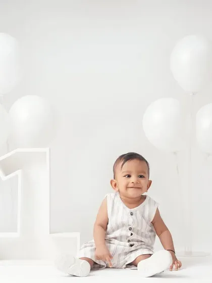 A simple and beautiful first birthday portrait of a baby boy in a minimalist all-white studio with white balloons and a large '1' prop.