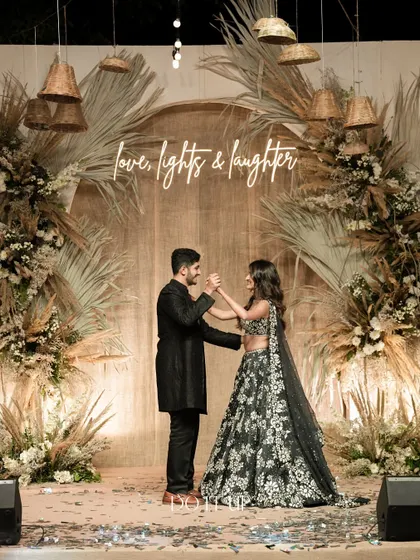 The couple shares a dance on the sangeet stage, which features a backdrop of dried palm leaves, pampas grass, and a custom neon sign. The design is a perfect blend of rustic and modern aesthetics.