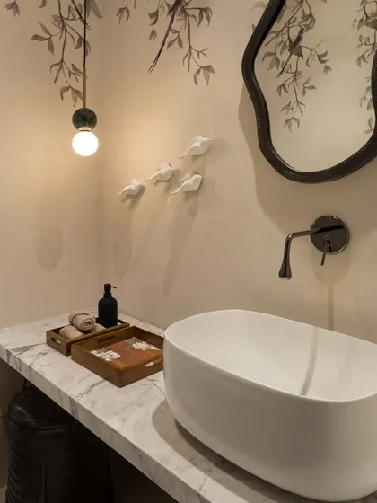 The powder room in the House of Terraces features a custom hand-painted wallpaper with a delicate bird and branch motif. The organic-shaped mirror and simple vessel sink complement the artistic, natural theme.