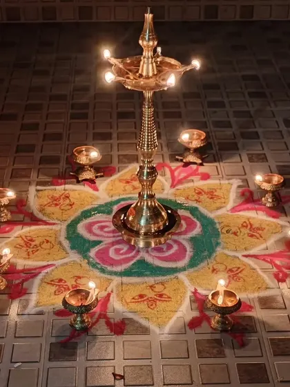 A traditional brass lamp, or 'Kuthu Vilakku', surrounded by small diyas on a colorful rangoli. This is how we celebrate Karthigai Deepam, the festival of lights.