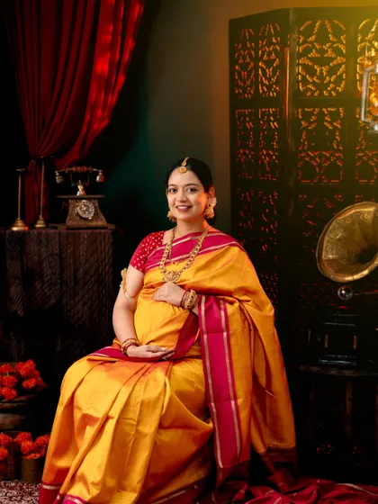 A classic vintage-themed maternity portrait. The mom-to-be is seated gracefully in a yellow and pink silk saree, surrounded by antique props like a gramophone and a classic telephone.