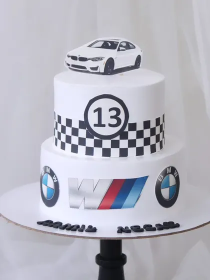 A sleek BMW-themed cake for a 13th birthday. This two-tiered cake features the iconic checkered racing pattern, BMW logos, and is topped with a model of a white BMW car.
