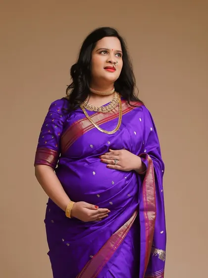 A classic studio portrait of a mother-to-be in a vibrant purple silk saree. The simple, clean background allows the rich color and texture of the fabric to stand out.