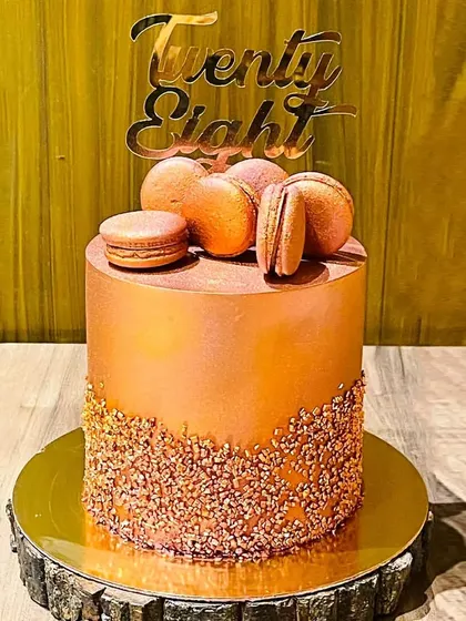 An elegant and modern 28th birthday cake. This custom chocolate walnut cake has a sleek metallic bronze finish, crunchy praline at the base, and is topped with delicate, handmade macarons.