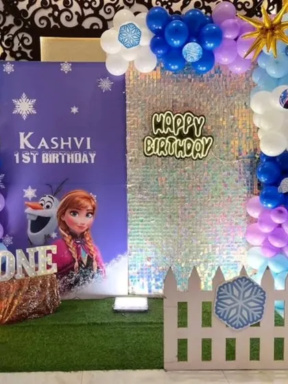 A first birthday celebration with a Frozen theme, featuring a shimmer wall, a purple backdrop with Anna and Olaf, and balloon columns in blue, purple, and white.