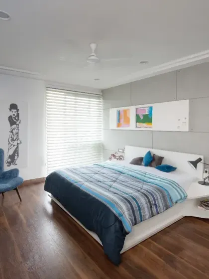 The master bedroom in the Grey Apartment is anchored by a sleek white platform bed and a textured grey accent wall. Rich wooden flooring and a plush blue accent chair add warmth and comfort.