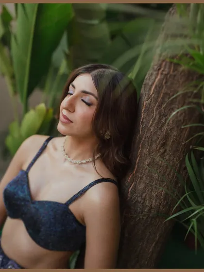 A serene portrait of a bride with her eyes closed, leaning against a tree trunk amidst lush foliage. This glamorous and peaceful shot highlights the beauty of the bride in a natural setting.