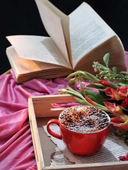 A cozy scene with a red coffee mug on a wooden tray, surrounded by flowers and an open book. The warm lighting and rich colors create a comfortable and inviting mood.