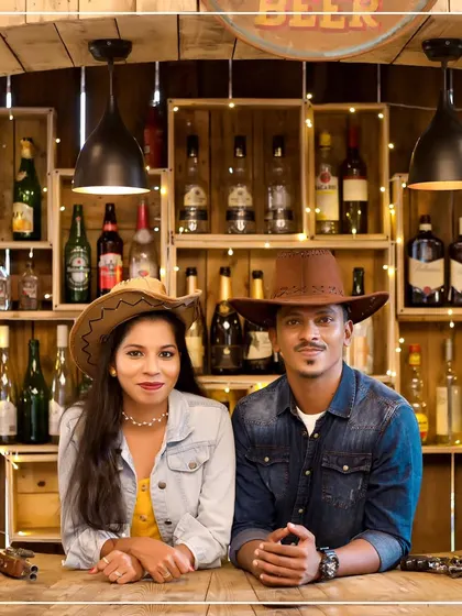 A themed pre-wedding shoot with a rustic, western vibe. The couple, dressed in cowboy hats and denim, poses at a wooden bar, creating a fun and memorable photograph that shows their unique style.
