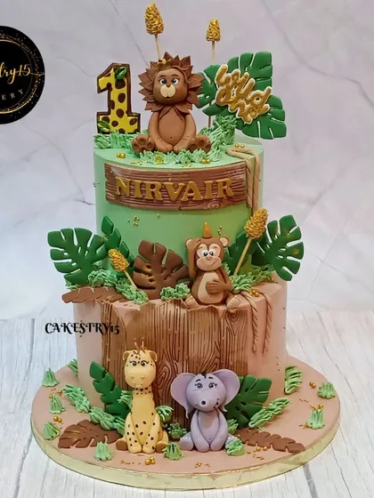 A wild two-tier jungle-themed cake for a first birthday. This cake is adorned with adorable fondant animals including a lion, monkey, giraffe, and elephant amidst lush green leaves.