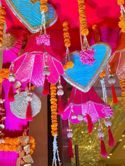 Intricate hanging decorations designed for a Garba night. This image shows custom-made fabric lampshades and heart-shaped ornaments in bright pink and blue, accented with marigold garlands and silver tassels.