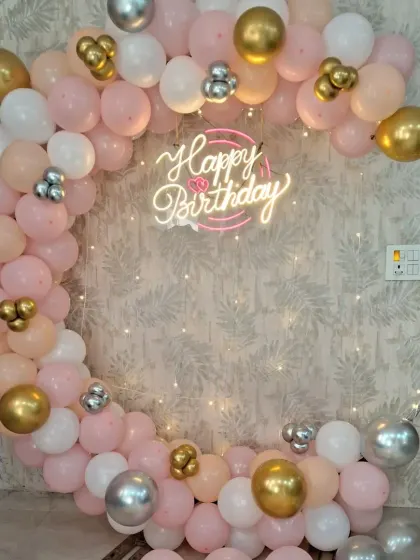 A beautiful birthday backdrop with a circular frame and a balloon garland in shades of pink, white, and gold. The 'Happy Birthday' neon sign adds a warm and celebratory glow.