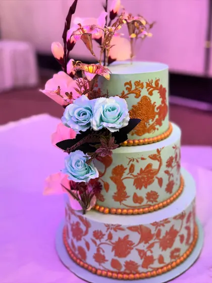 An elegant three-tier wedding cake with a mint green base and intricate gold stenciling. The design is completed with a cascade of delicate blue and pink sugar flowers.