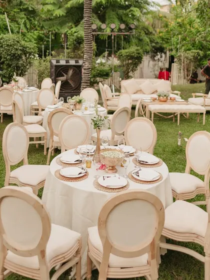 An elegant daytime setup for a 75th anniversary in a garden. The neutral color palette with cream-colored linens and classic wooden chairs creates a timeless and sophisticated outdoor luncheon feel.