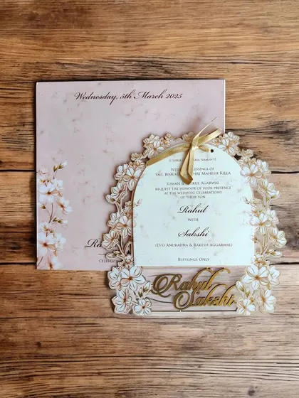 A delicate and romantic MDF laser-cut invitation with a floral arch frame. The pastel pink background and gold ribbon add to its charm.