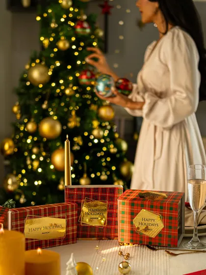 A scene of a woman decorating a Christmas tree, with gift boxes elegantly placed on a table in the foreground. This tells a story of holiday preparation.
