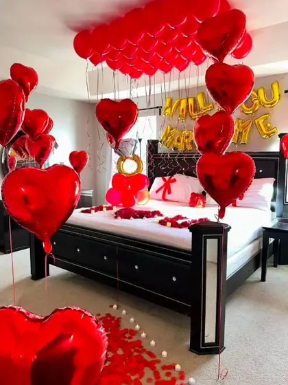 A grand proposal room setup filled with red heart-shaped foil balloons. The decor includes 'Will You Marry Me' foil balloons over the bed and a large engagement ring balloon arrangement.