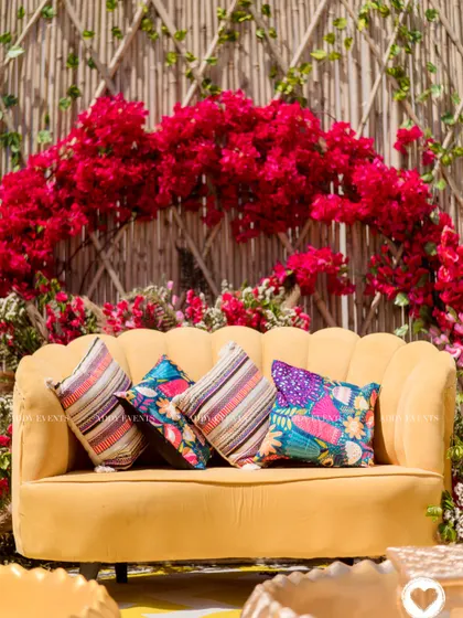A close-up of the vibrant Haldi seating, where a mustard yellow sofa is set against a dramatic arch of red bougainvillea and a bamboo lattice wall.