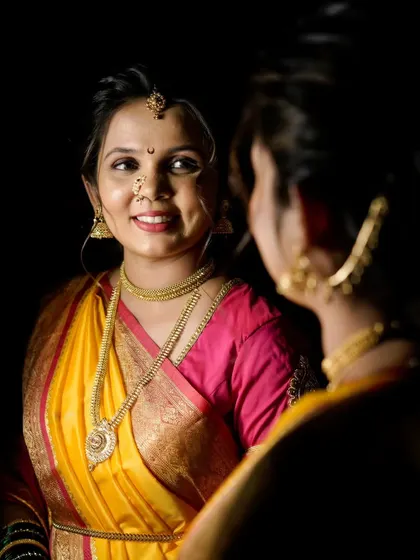 A classic mirror shot, capturing the bride's reflection as she gets ready. The dark background makes her yellow and pink saree pop, highlighting her happy anticipation.