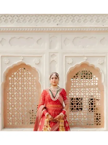A full-length portrait of the bride standing before a traditional jali wall. Her regal pose and stunning red lehenga make for a powerful and elegant bridal photograph.