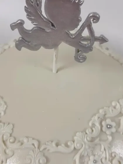 A close-up of an elegant, square-shaped cake with intricate white lace piping and a silver angel topper.