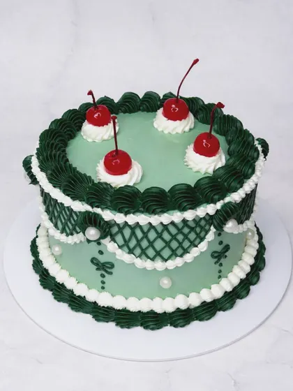 We're obsessed with the charm of vintage Lambeth cakes, and this one is a favorite. The intricate piping in forest green and white, topped with classic maraschino cherries, shows our dedication to this beautiful, ornate cake decorating style.